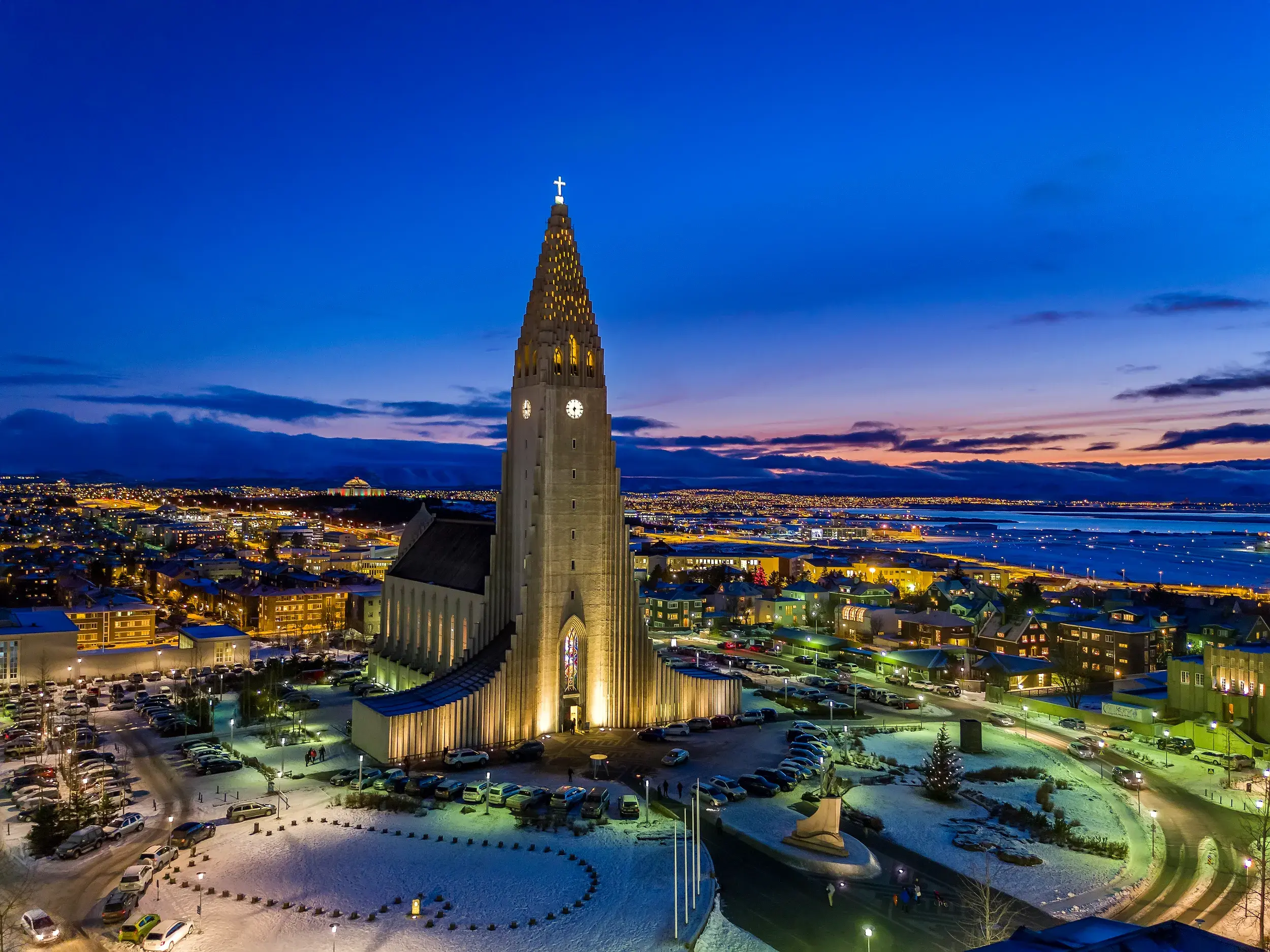 Die beleuchtete Hallgrimskirche bei Dämmerung, umgeben vom winterlichen Reykjavik mit seinen Lichtern, die bis zum Meer reichen.