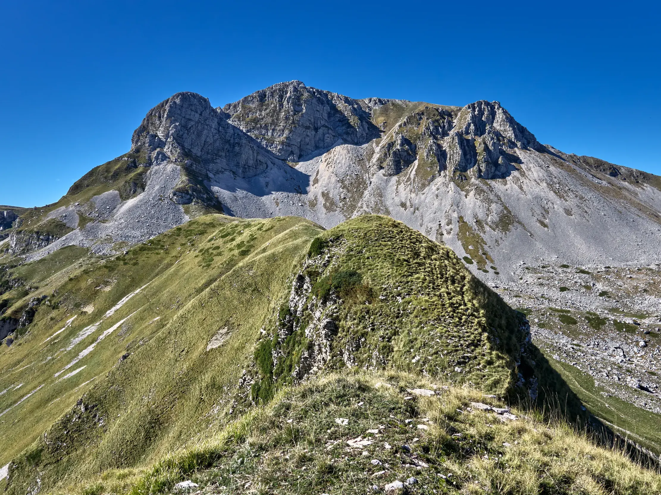 Ein grüner Bergpfad führt über einen Kamm zum felsigen Gipfel des Monte Meta im Nationalpark der Abruzzen bei Sonnenschein.