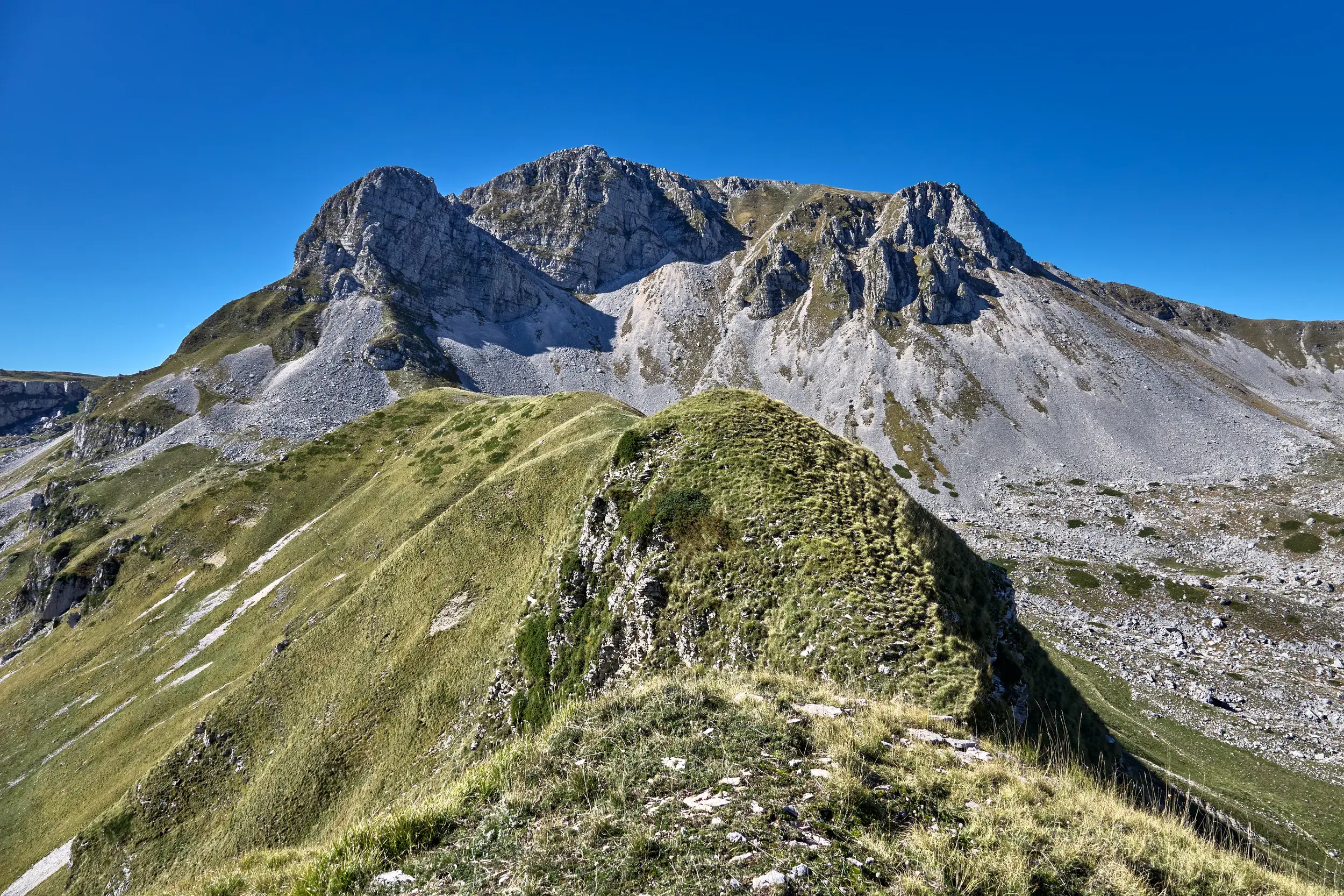 Ein grüner Bergpfad führt über einen Kamm zum felsigen Gipfel des Monte Meta im Nationalpark der Abruzzen bei Sonnenschein.