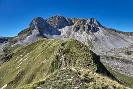 Ein grüner Bergpfad führt über einen Kamm zum felsigen Gipfel des Monte Meta im Nationalpark der Abruzzen bei Sonnenschein.