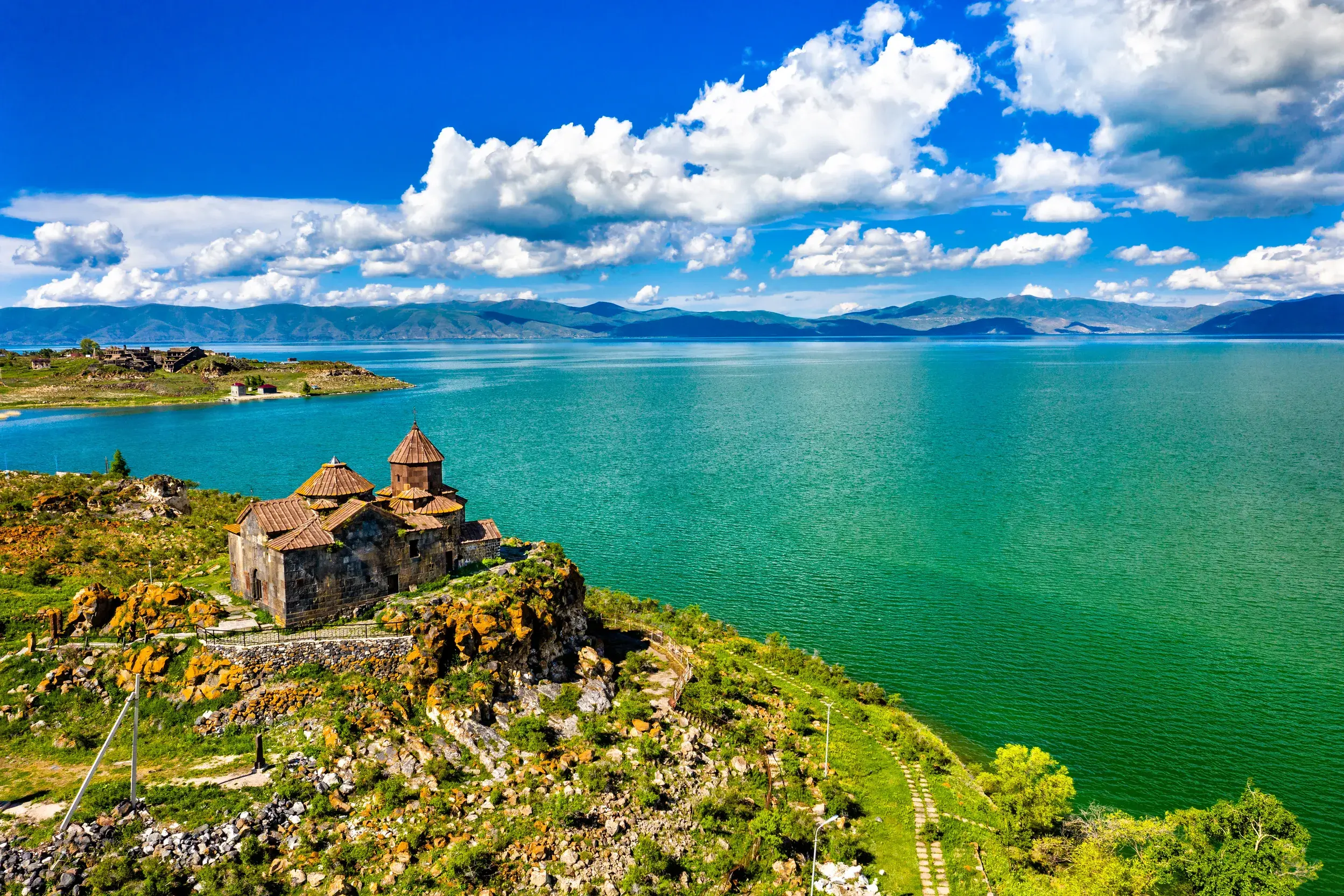 Historisches Sewankloster auf einer grünen Halbinsel am türkisfarbenen Sewansee in Armenien vor einer Bergkulisse.