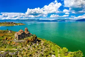 Historisches Sewankloster auf einer grünen Halbinsel am türkisfarbenen Sewansee in Armenien vor einer Bergkulisse.
