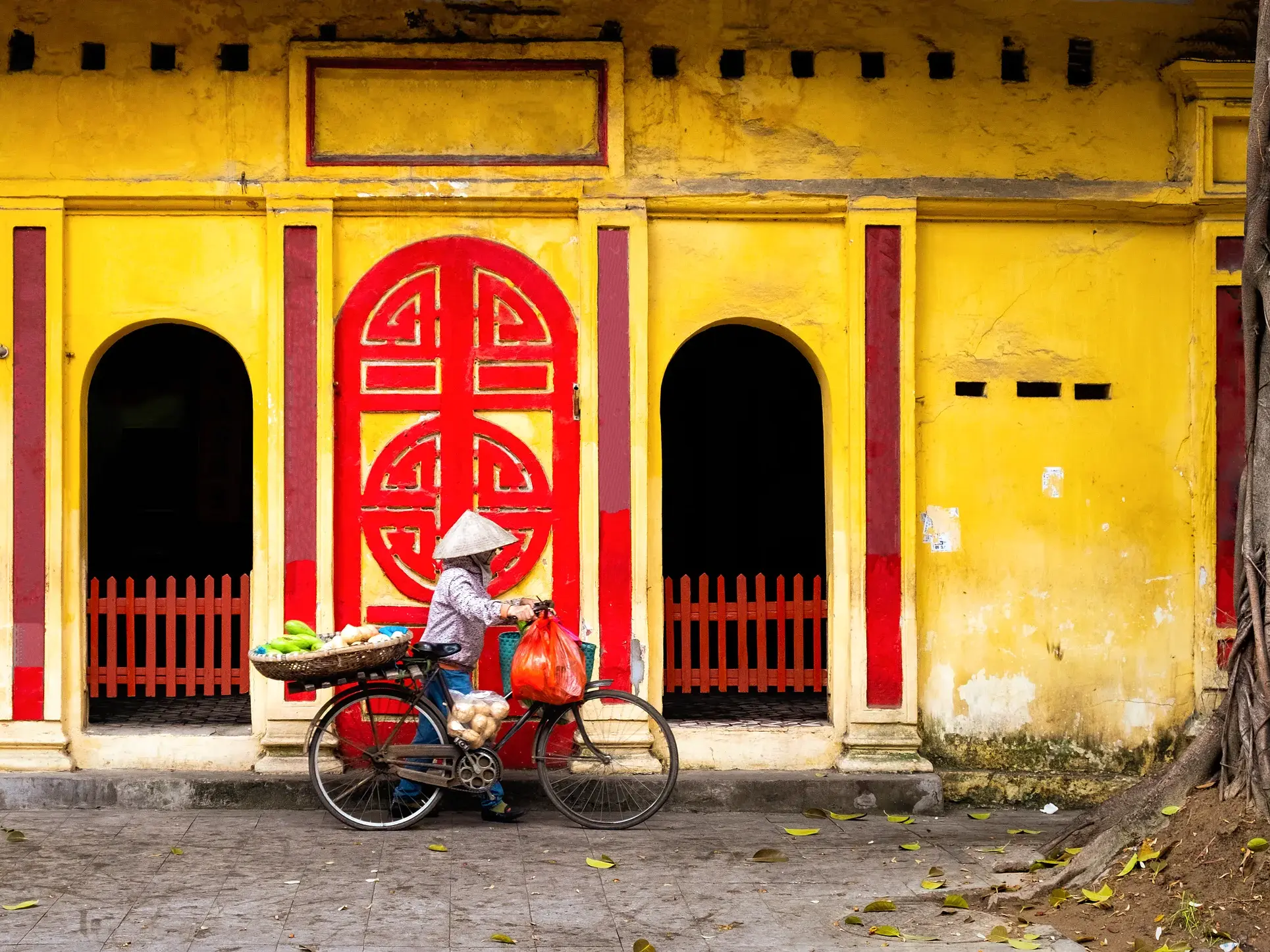Eine Händlerin mit Kegelhut schiebt ihr Fahrrad an einer leuchtend gelben Hausfassade in der Altstadt von Hanoi entlang.