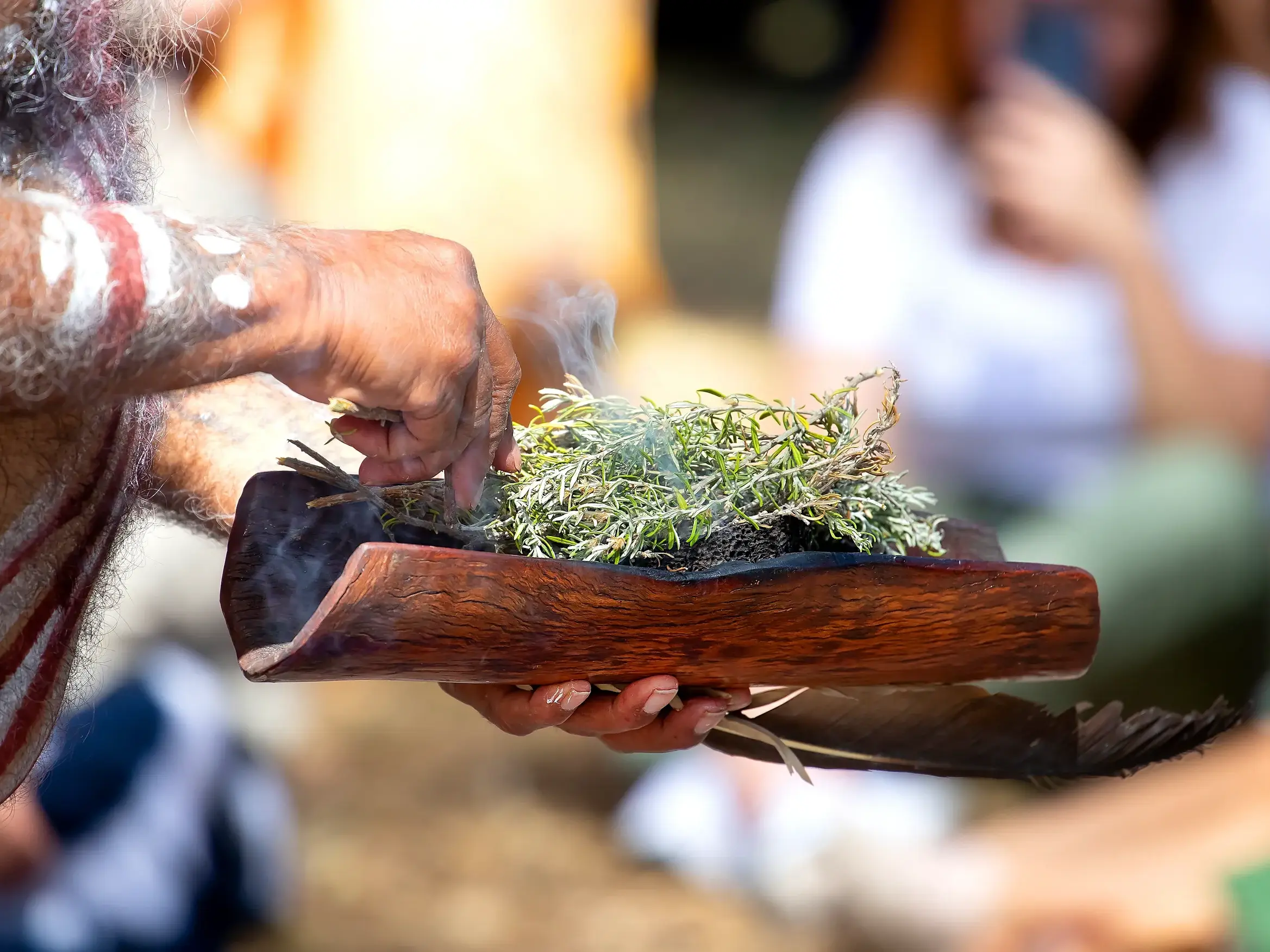 Ein australischer Ureinwohner mit Körperbemalung führt eine traditionelle Rauchzeremonie mit Kräutern in einer Holzschale durch.