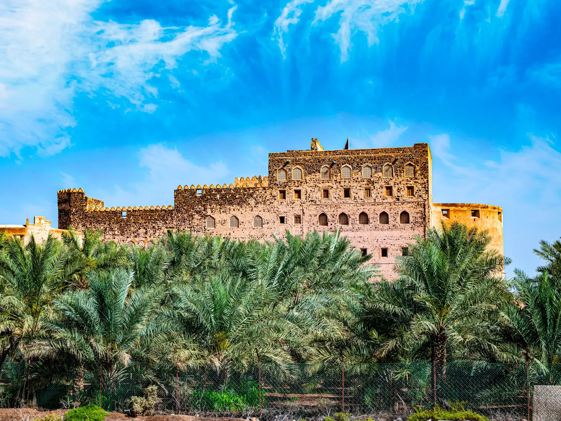 Die historische Festung Jabrin im Oman erhebt sich hinter grünen Palmenwipfeln unter einem strahlend blauen Himmel.