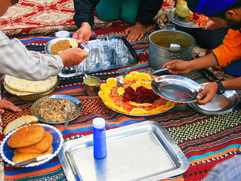 Eine Wandergruppe teilt sich mit marokkanischen Begleitern ein Picknick auf einer bunten Decke im Hohen Atlas.