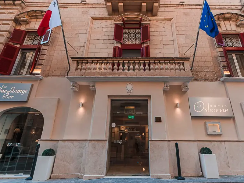 Die historische Steinfassade des Hotel Osborne in Valletta mit dem gläsernen Eingang, Balkon und maltesischer sowie europäischer Flagge.