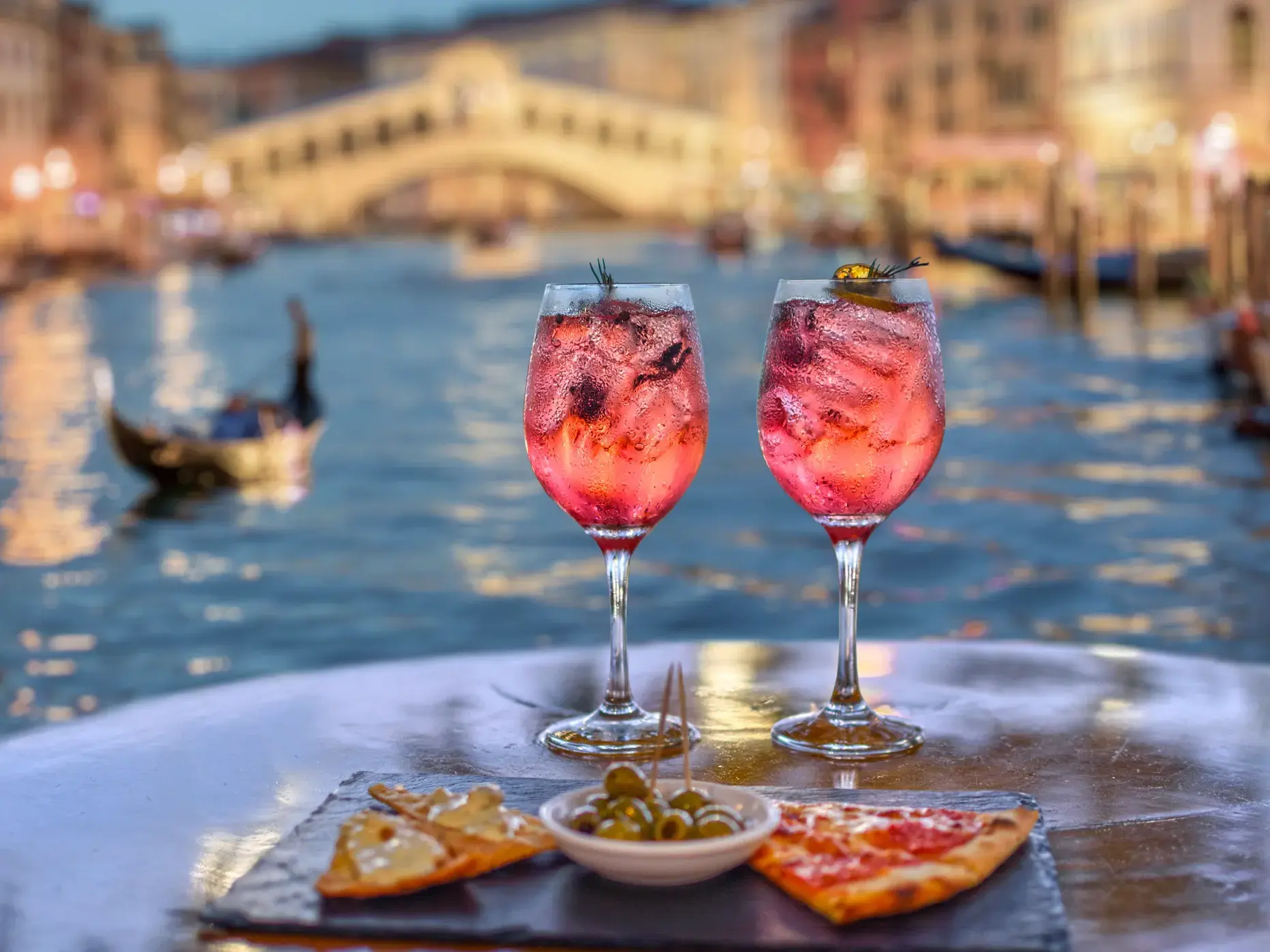 Aperitivo mit zwei Gläsern Sprizz und Blick auf den Canal Grande mit der Rialtobrücke in Venedig bei Dämmerung.