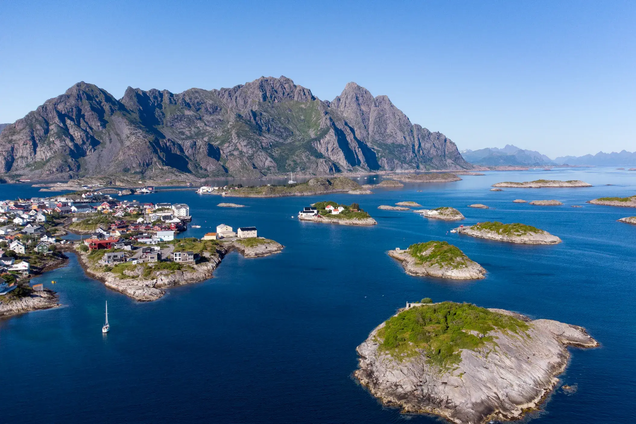 Luftaufnahme des Fischerdorfs Henningsvaer auf den Lofoten, umgeben von der blauen See und Felseninseln am Fuße der Berge.