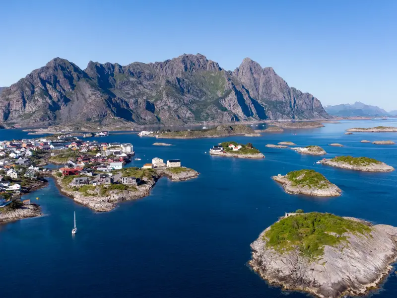 Luftaufnahme des Fischerdorfs Henningsvaer auf den Lofoten, umgeben von der blauen See und Felseninseln am Fuße der Berge.