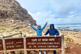Eine Frau und ein Mann posieren fröhlich am Schild des Kaps der Guten Hoffnung vor rauer Felsküste und dem Meer.