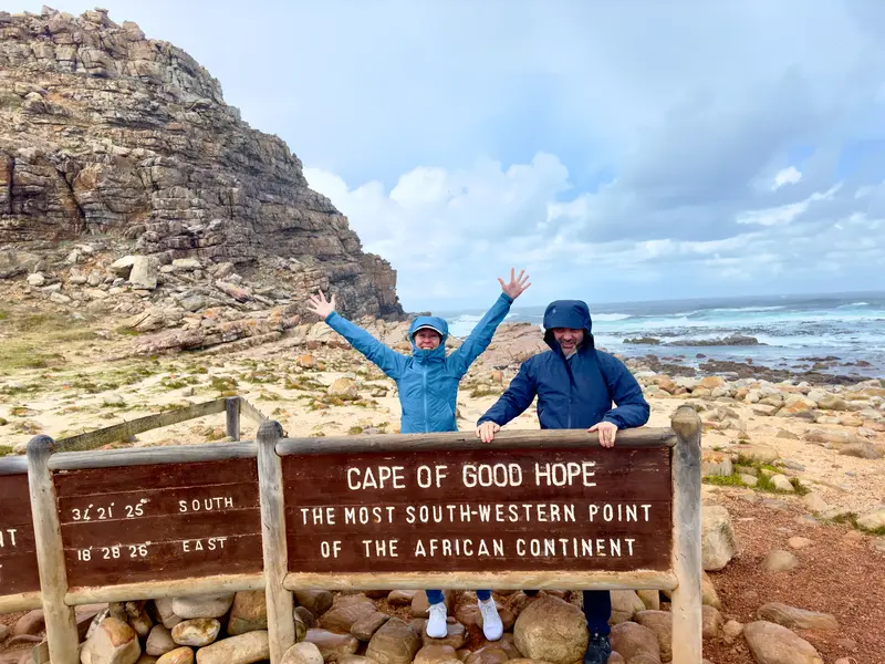 Eine Frau und ein Mann posieren fröhlich am Schild des Kaps der Guten Hoffnung vor rauer Felsküste und dem Meer.