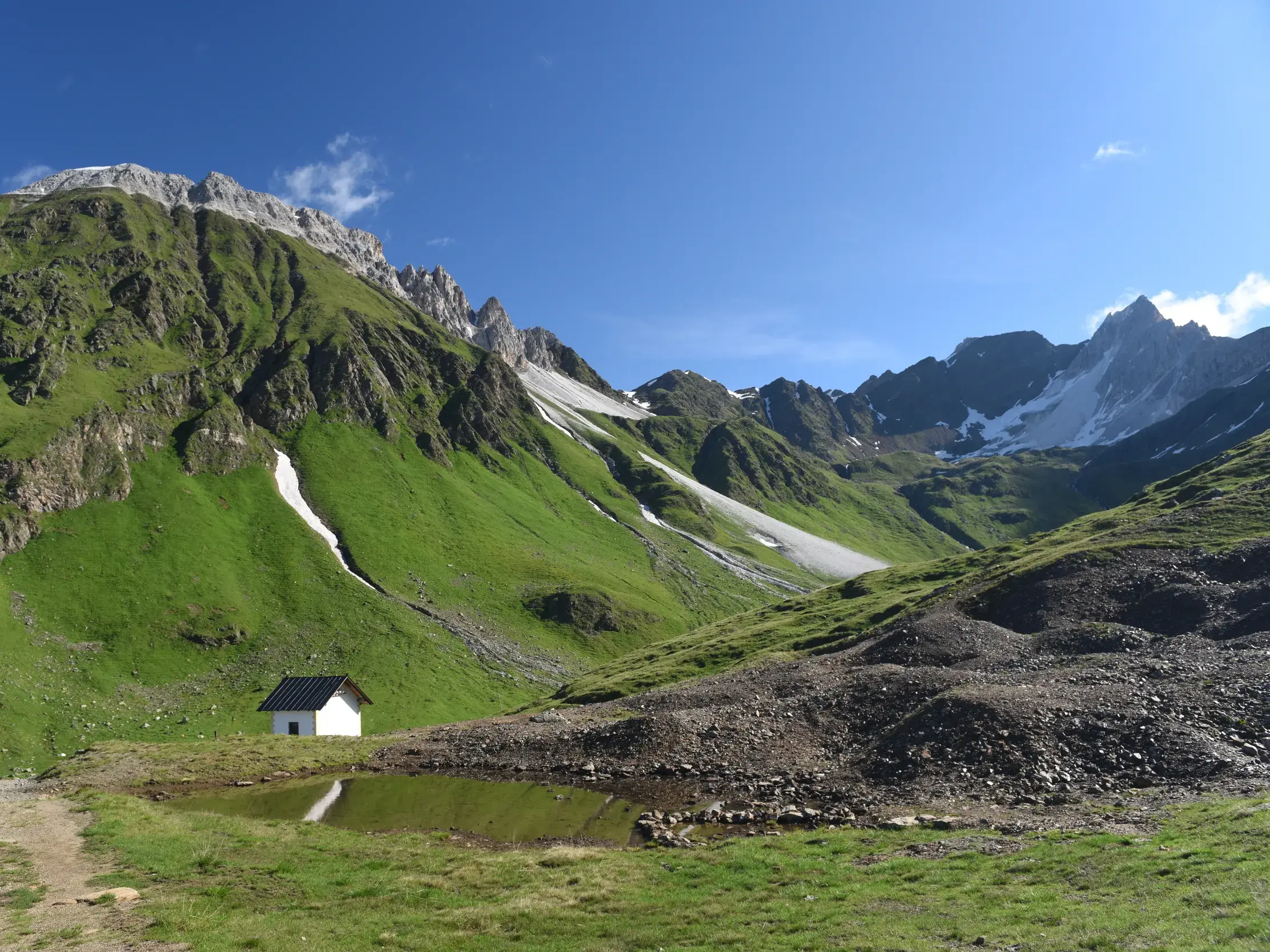 Eine einsame, weiße Berghütte an einem kleinen Teich in einem weiten, grünen Tal der Alpen unter blauem Himmel.