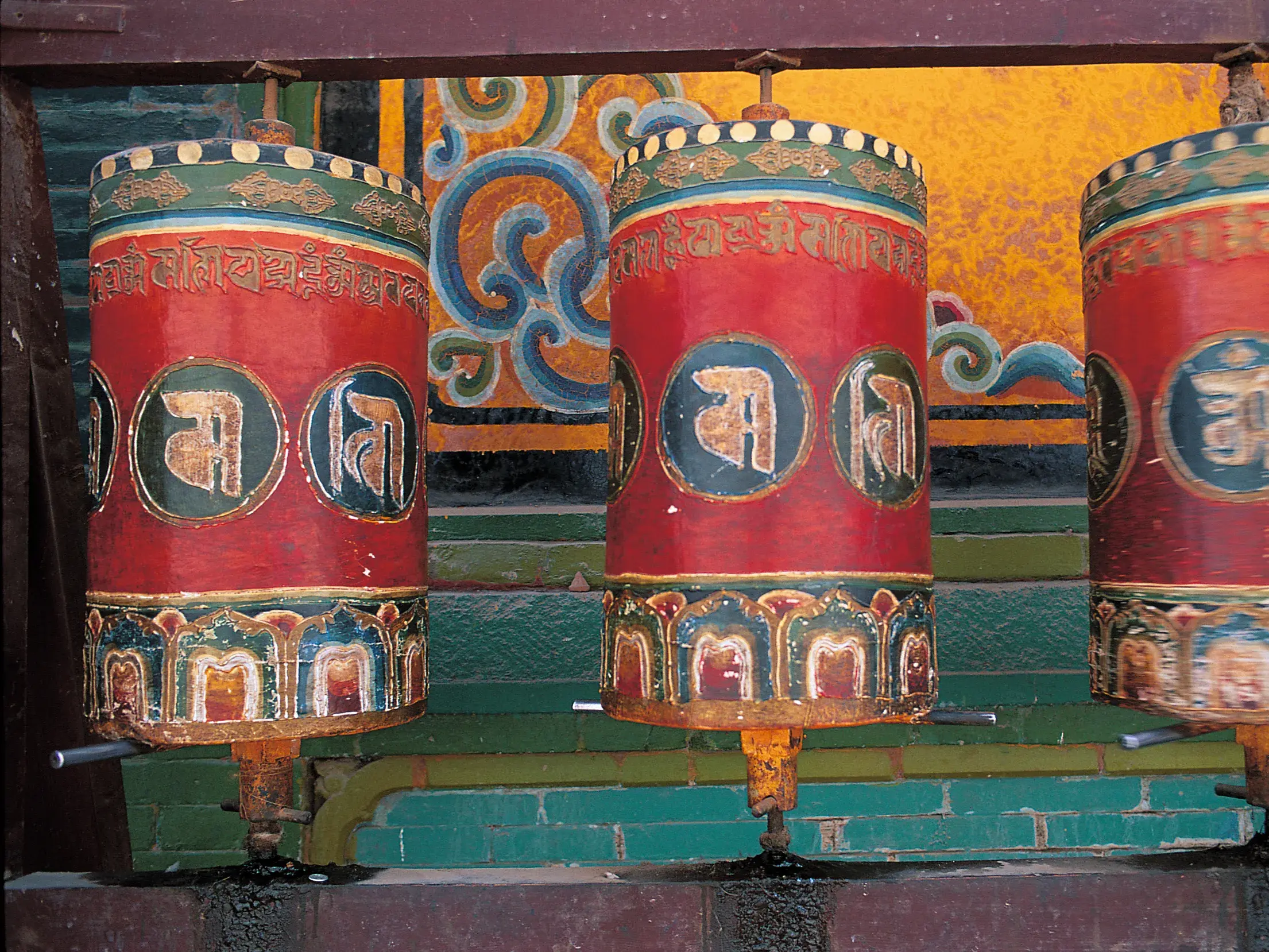 Nahaufnahme von drei roten, kunstvoll bemalten Gebetsmühlen mit tibetischen Schriftzeichen in einem Kloster in Tibet.