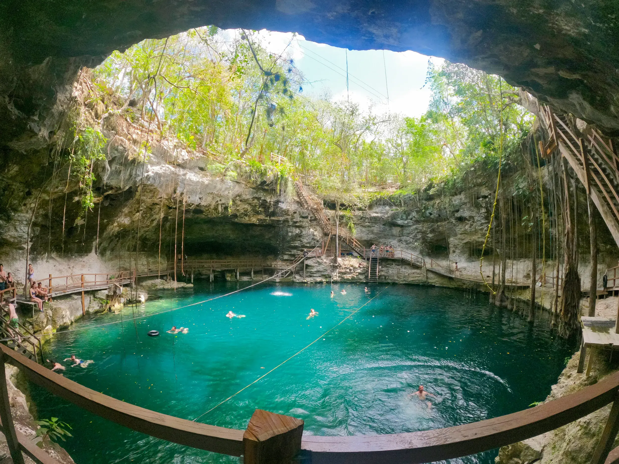 Reisende schwimmen im kristallklaren Wasser des Felsenpools Cenote Xcanché in Mexiko, während Lianen von der Höhlendecke hängen.