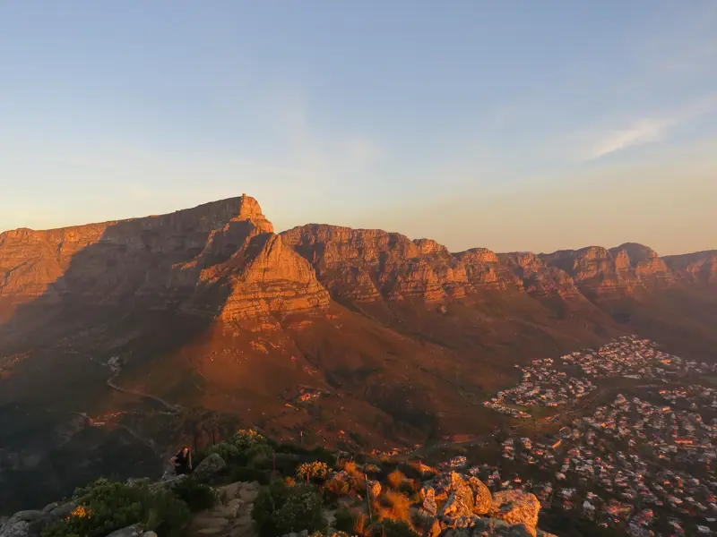 Panoramablick auf den Tafelberg bei Sonnenaufgang mit einem Wanderer im Vordergrund und Kapstadt am Fuße des Berges.