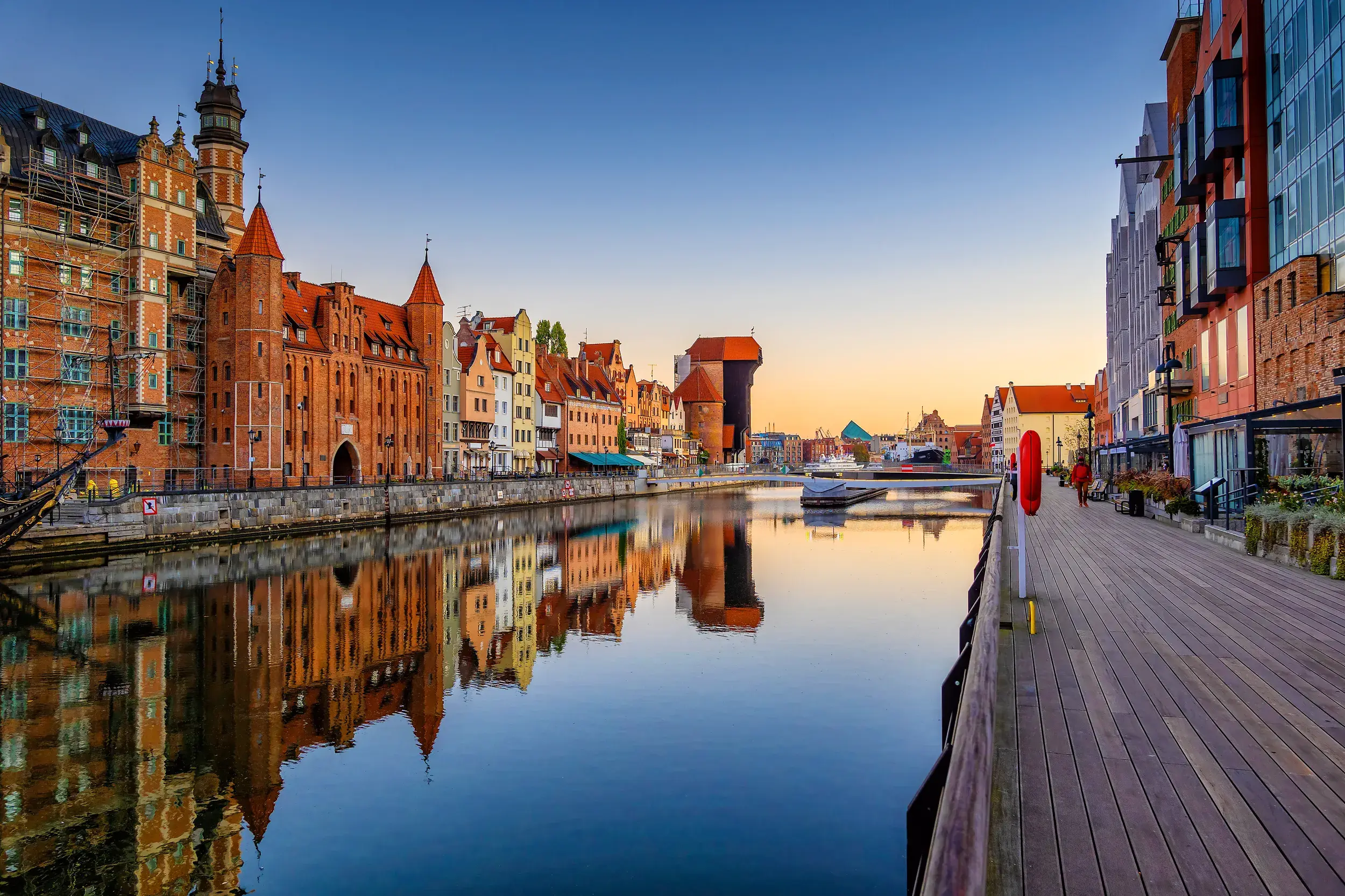 Historische Patrizierhäuser und das Krantor in Danzig spiegeln sich bei Sonnenuntergang im Wasser der Uferpromenade.