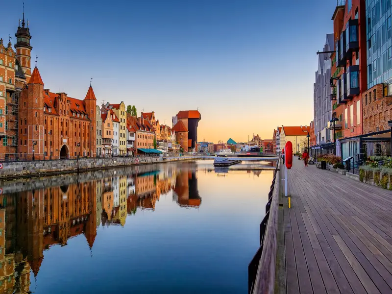 Historische Patrizierhäuser und das Krantor in Danzig spiegeln sich bei Sonnenuntergang im Wasser der Uferpromenade.