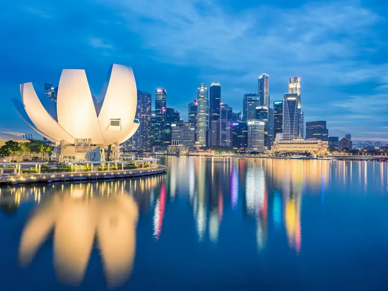 Das beleuchtete ArtScience Museum und die Skyline von Singapur spiegeln sich bei Dämmerung im ruhigen Wasser der Marina Bay.