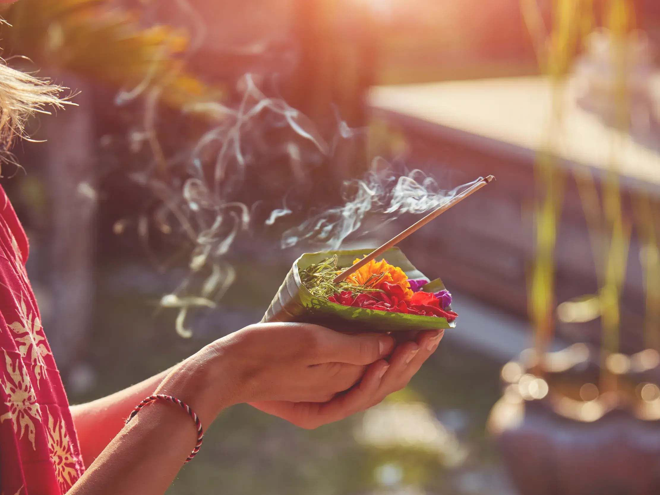Hände halten in Bali eine traditionelle Opfergabe mit bunten Blüten und einem rauchenden Räucherstäbchen in der Abendsonne.