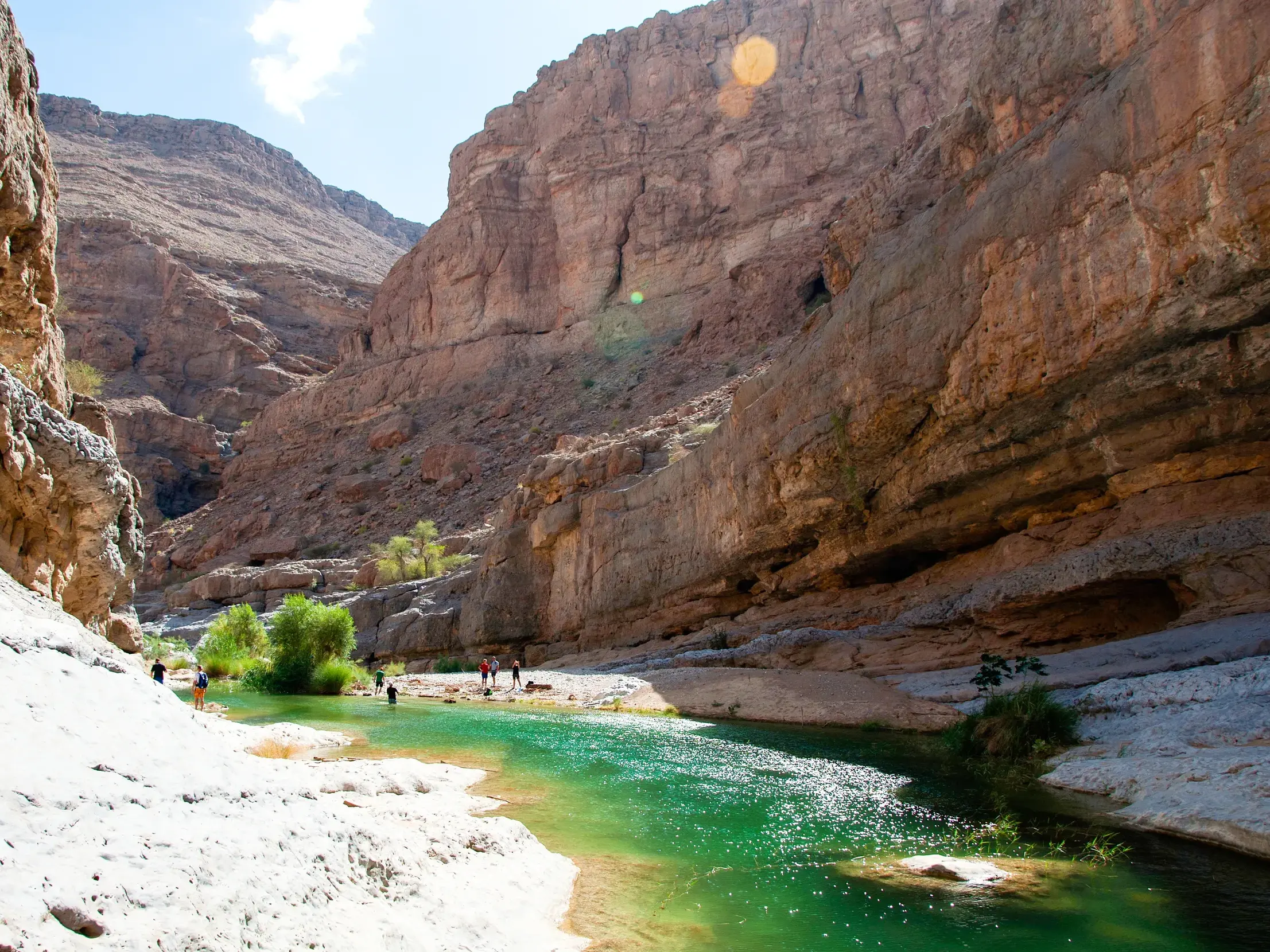 Wanderer einer Trekkinggruppe erkunden ein Wadi im Hadschar-Gebirge im Oman mit smaragdgrünem Wasser und steilen Felswänden.