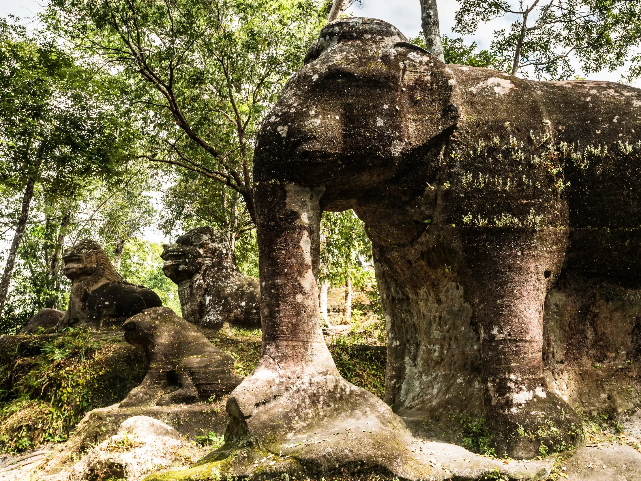 Große, moosbewachsene Elefanten-Skulptur aus Sandstein neben Löwenfiguren im Dschungel des Phnom Kulen Nationalparks in Kambodscha.
