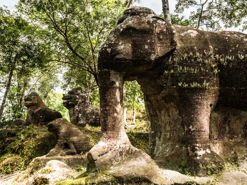 Große, moosbewachsene Elefanten-Skulptur aus Sandstein neben Löwenfiguren im Dschungel des Phnom Kulen Nationalparks in Kambodscha.
