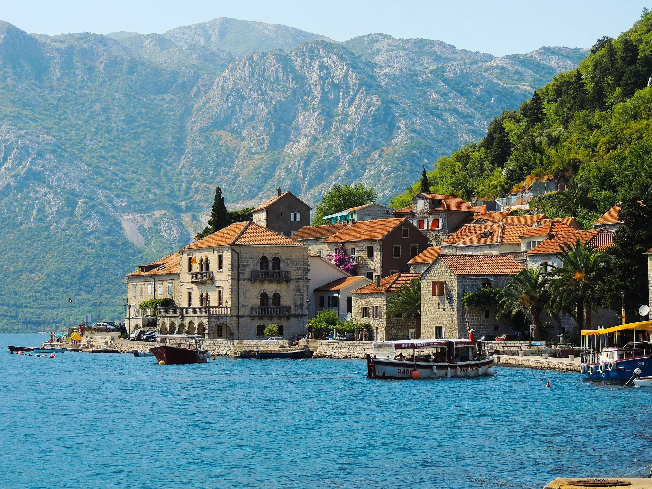 Ein Ausflugsboot fährt entlang der malerischen Küste von Perast in der Bucht von Kotor vor einer beeindruckenden Bergkulisse.