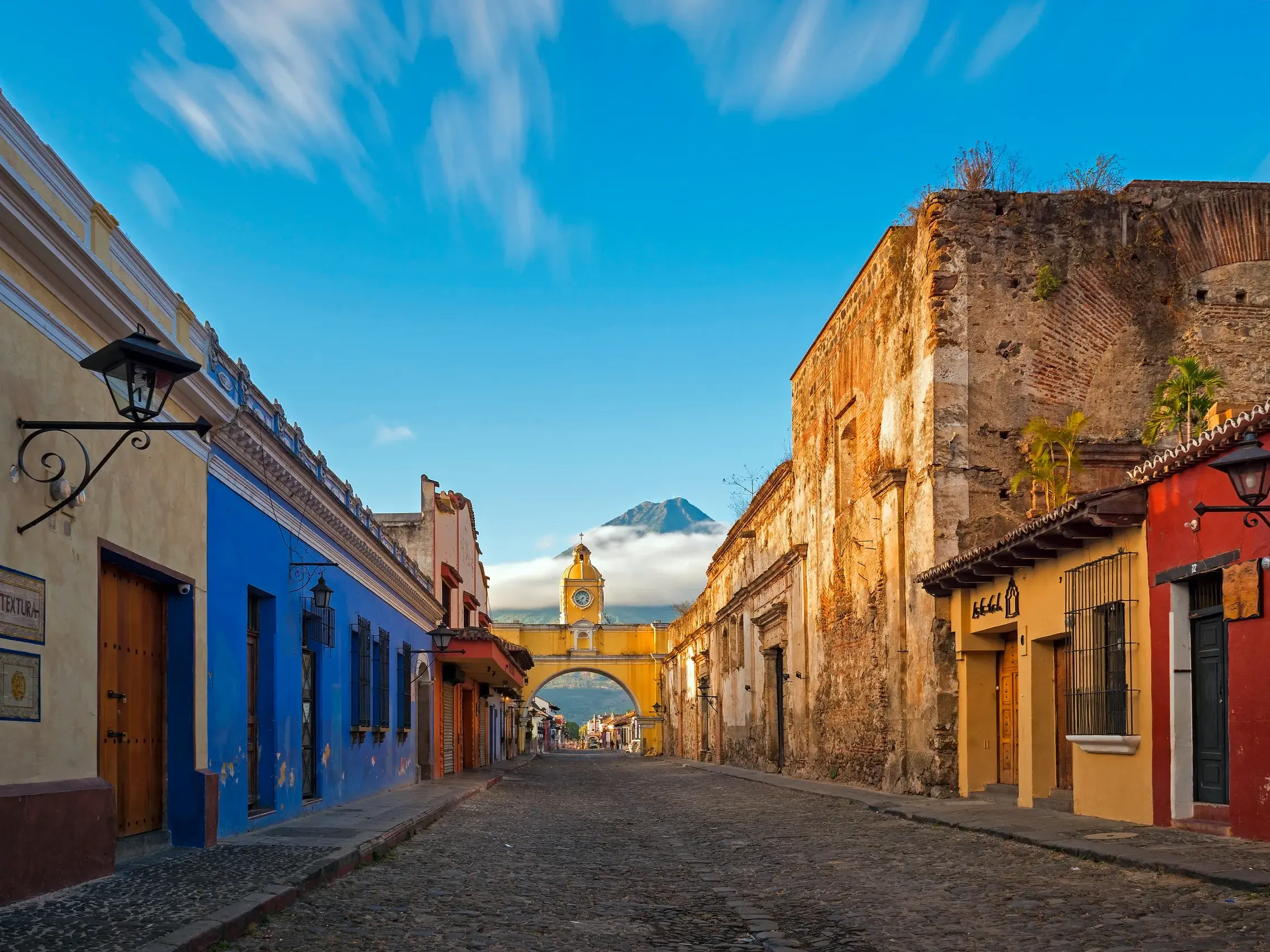 Blick durch eine Kopfsteinpflaster-Gasse in Antigua auf den gelben Bogen Santa Catalina mit dem Vulkan de Agua im Hintergrund.