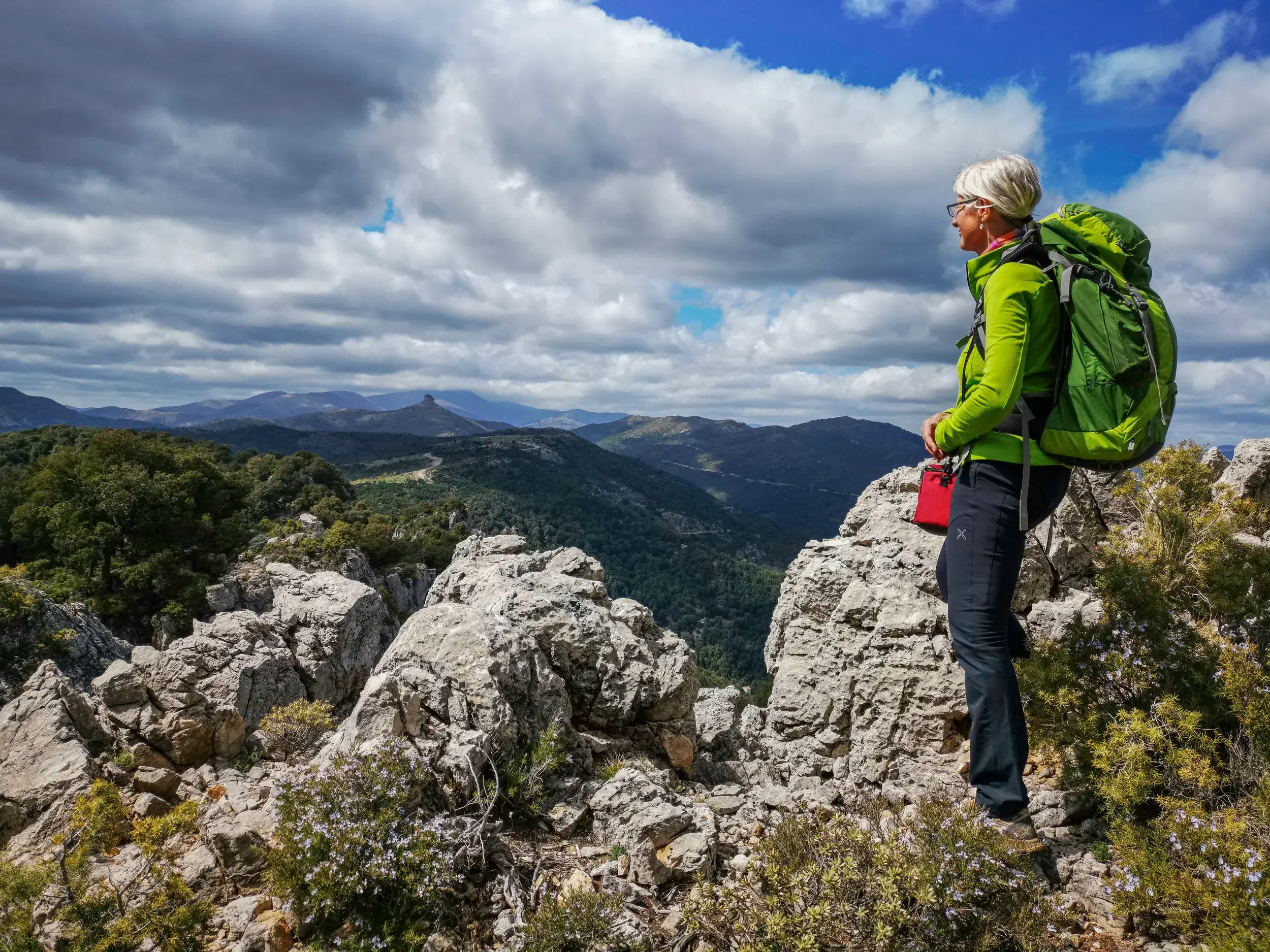 Eine Wanderin mit Rucksack blickt von einem Felsen auf die Berglandschaft Sardiniens mit dem Felsturm Perda Liana am Horizont.