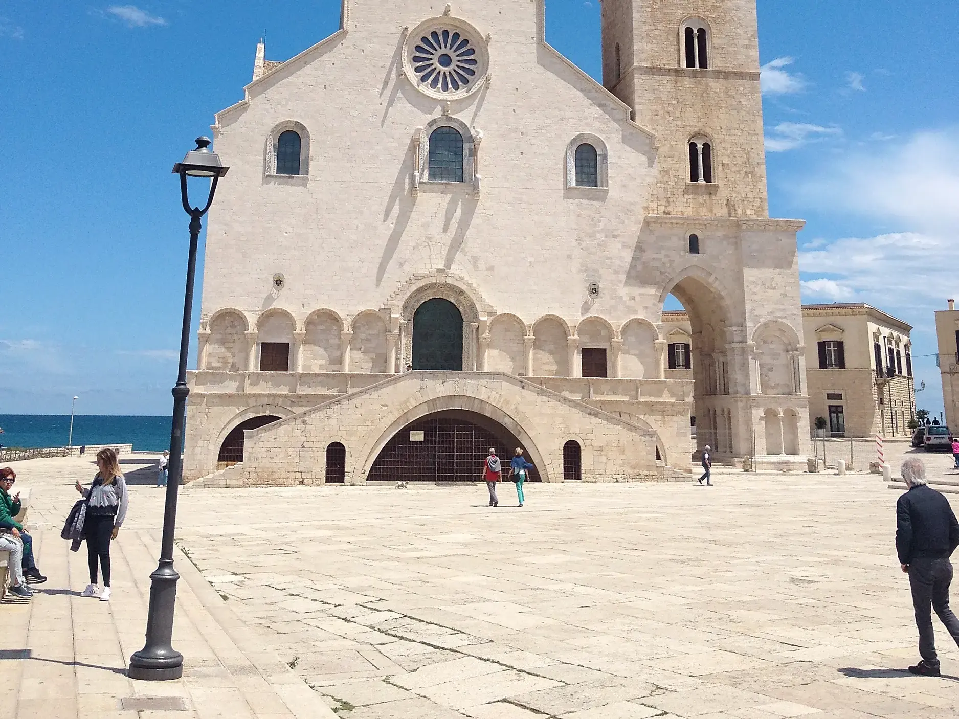 Die schneeweiße Kathedrale von Trani erhebt sich auf einem großen Platz direkt am Meer, Passanten flanieren in der Sonne.