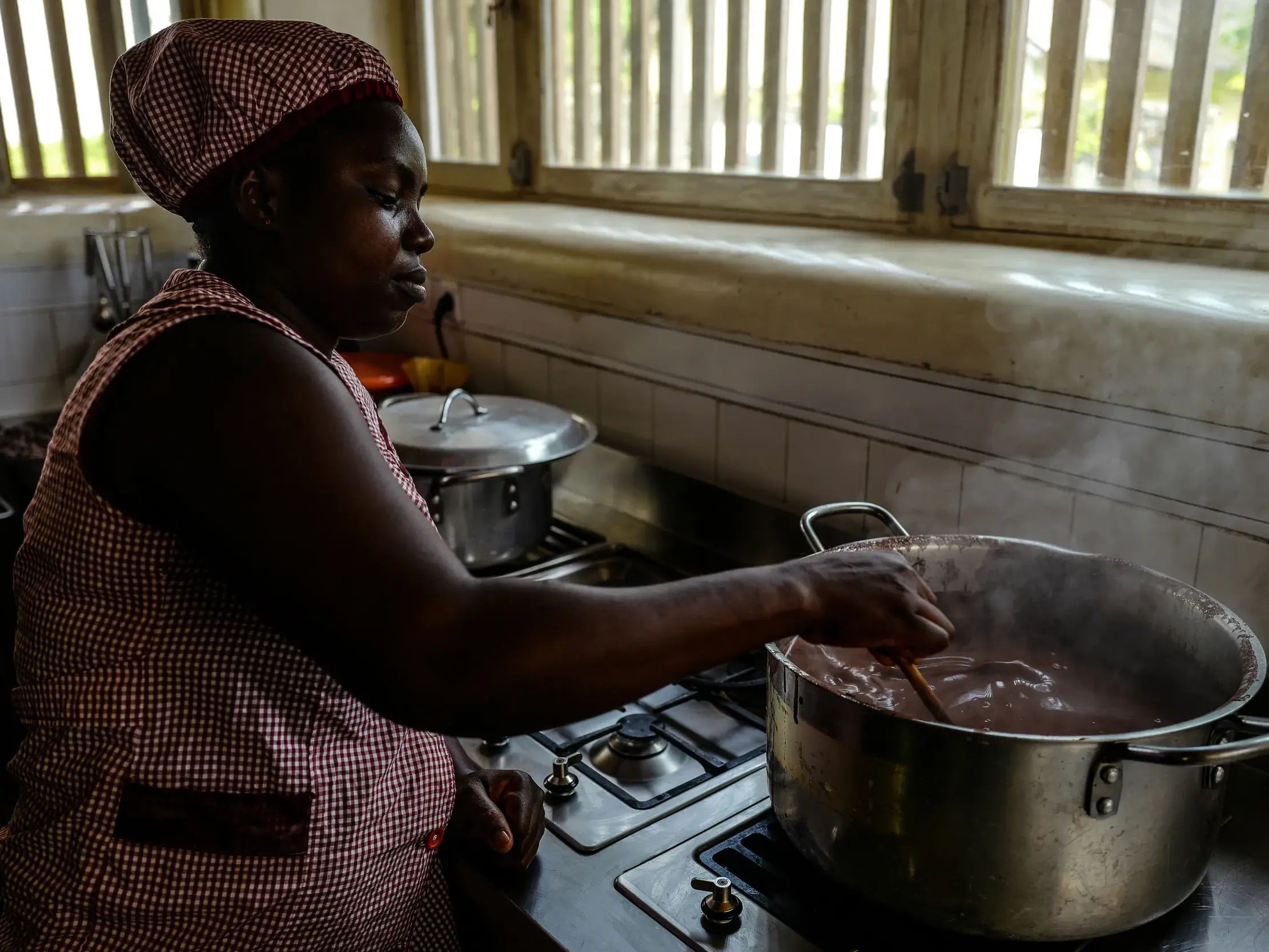Eine santomeische Frau in einer traditionellen Küche auf São Tomé rührt mit einem Holzlöffel in einem großen, dampfenden Topf.