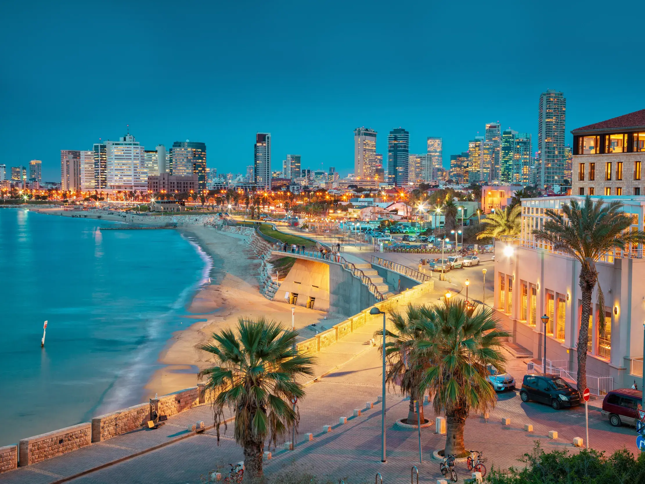 Abendlicher Blick auf Tel Avivs beleuchtete Skyline, die Strandpromenade und den Sandstrand am Mittelmeer bei Dämmerung.