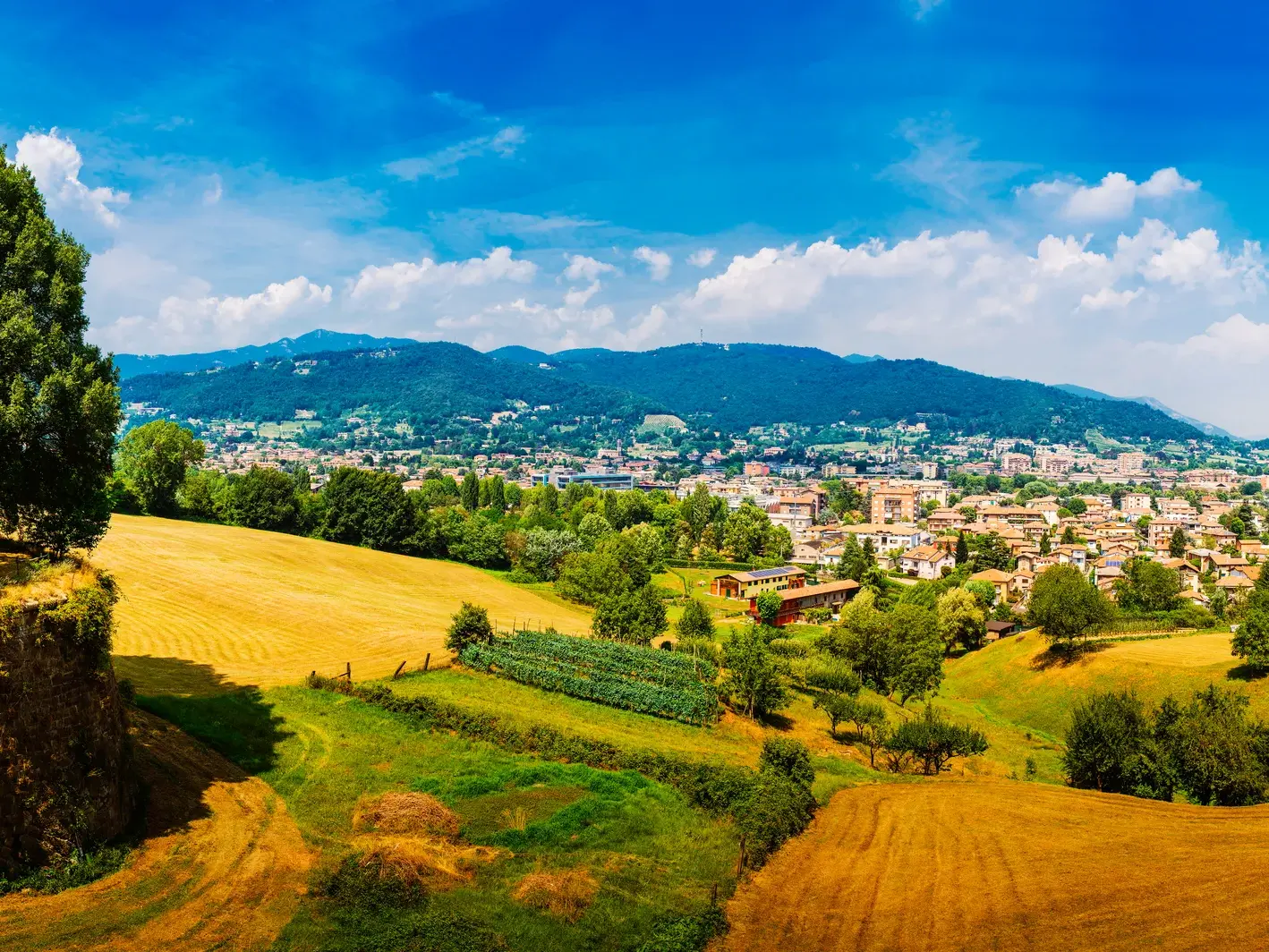 Panoramablick auf die Stadt Bergamo in der Lombardei, umgeben von grünen Hügeln und goldenen Feldern unter blauem Himmel.