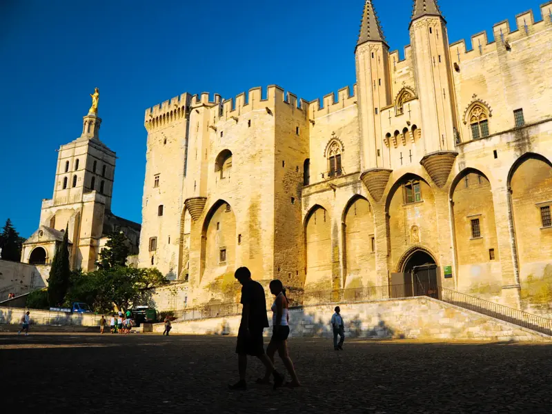 Besucher spazieren im warmen Abendlicht über den Vorplatz des monumentalen Papstpalastes in Avignon, einem UNESCO-Welterbe.