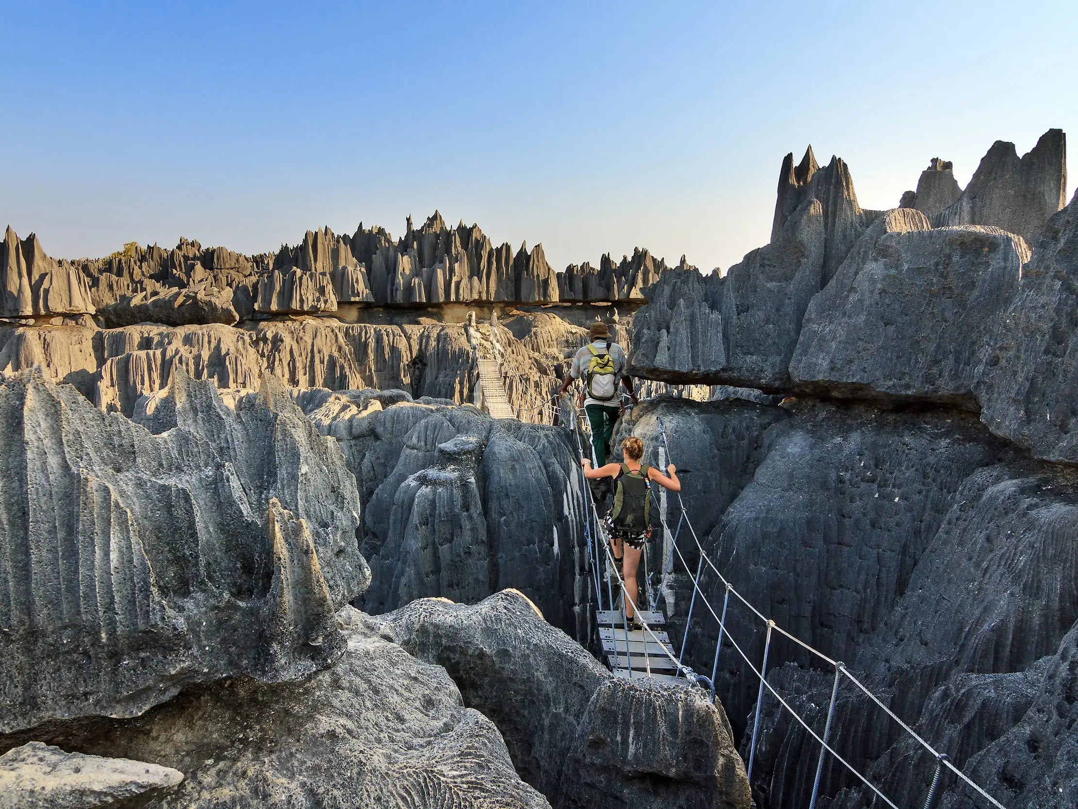 Zwei Wanderer auf einer Hängebrücke zwischen spitzen Kalksteinfelsen im Nationalpark Tsingys de Bemaraha in Madagaskar.