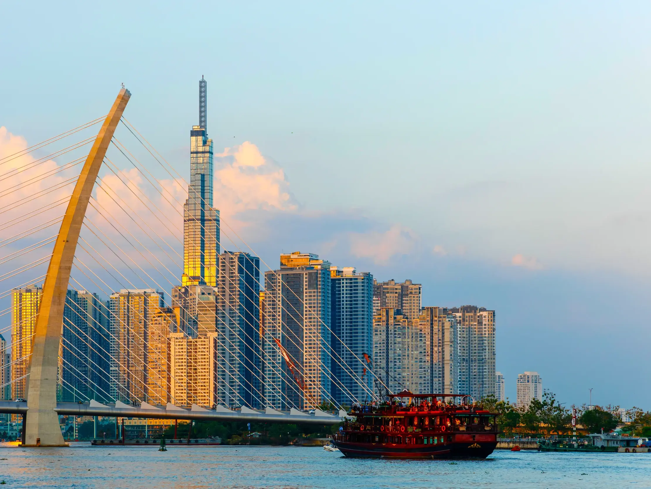 Ein rotes Ausflugsboot auf dem Saigon-Fluss vor der modernen Skyline von Ho-Chi-Minh-Stadt mit dem Landmark 81 im Abendlicht.