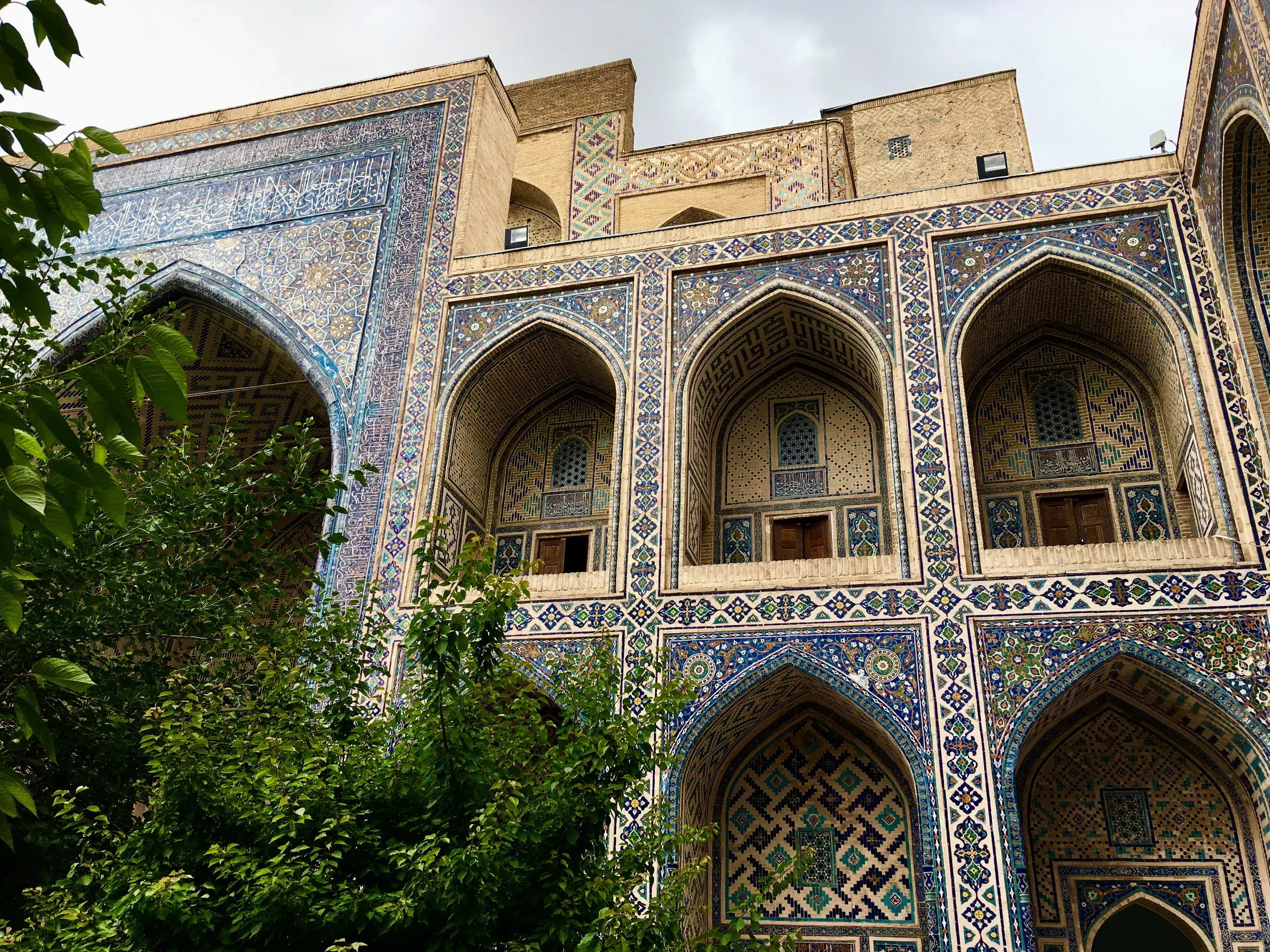 Kunstvolle Mosaikfassade einer Medrese in Samarkand mit Spitzbögen und blau-türkisen Fliesenmustern, gesehen hinter grünen Bäumen.