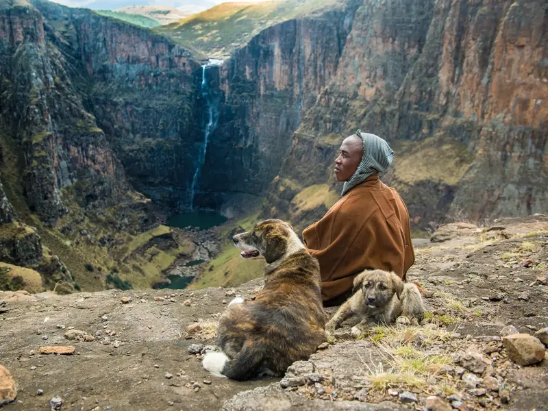 Einheimischer sitzt mit zwei Hunden auf einem Felsvorsprung und blickt in eine tiefe Schlucht der Drakensberge in Südafrika.