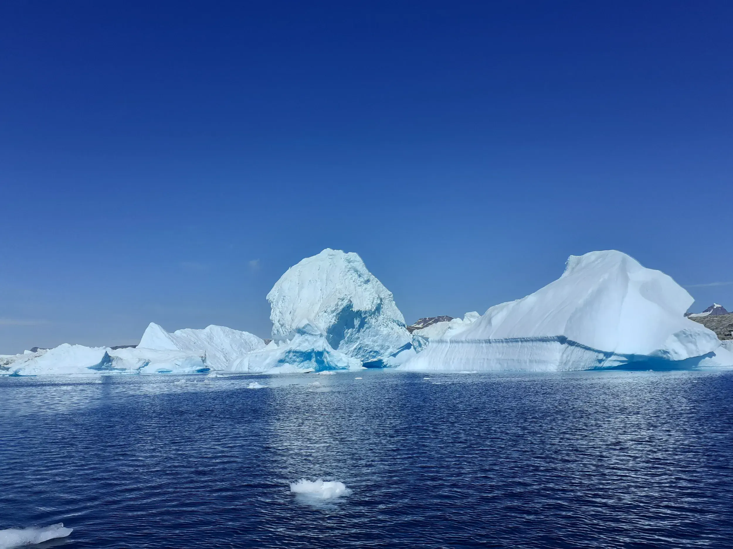 Majestätische Eisberge treiben im tiefblauen Wasser des Sermilik-Fjords in Ostgrönland unter klarem, sonnigem Himmel.