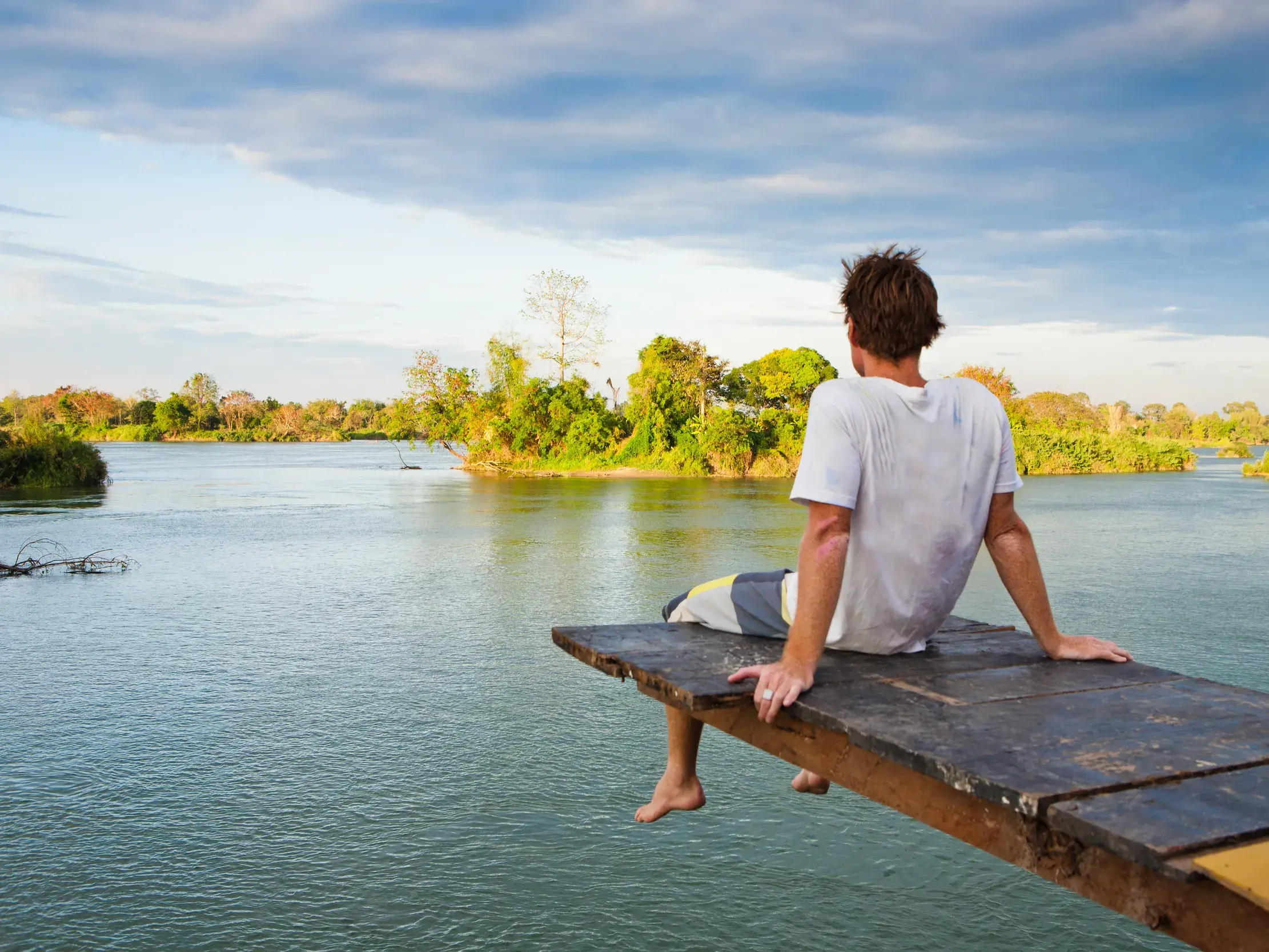 Ein Mann entspannt auf einem Holzsteg über dem Mekong und blickt auf die üppig grüne Flusslandschaft der 4000 Inseln in Laos.