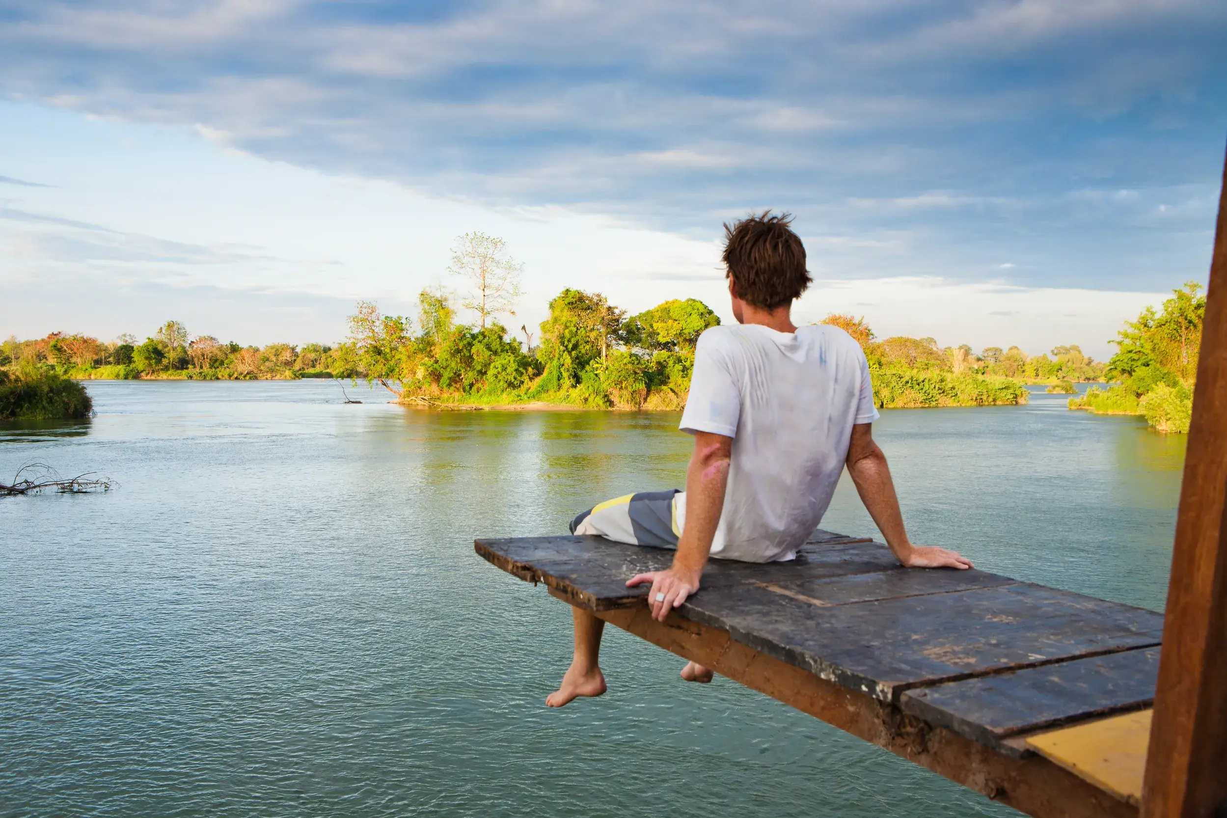 Ein Mann entspannt auf einem Holzsteg über dem Mekong und blickt auf die üppig grüne Flusslandschaft der 4000 Inseln in Laos.