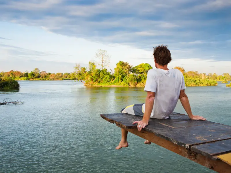 Ein Mann entspannt auf einem Holzsteg über dem Mekong und blickt auf die üppig grüne Flusslandschaft der 4000 Inseln in Laos.