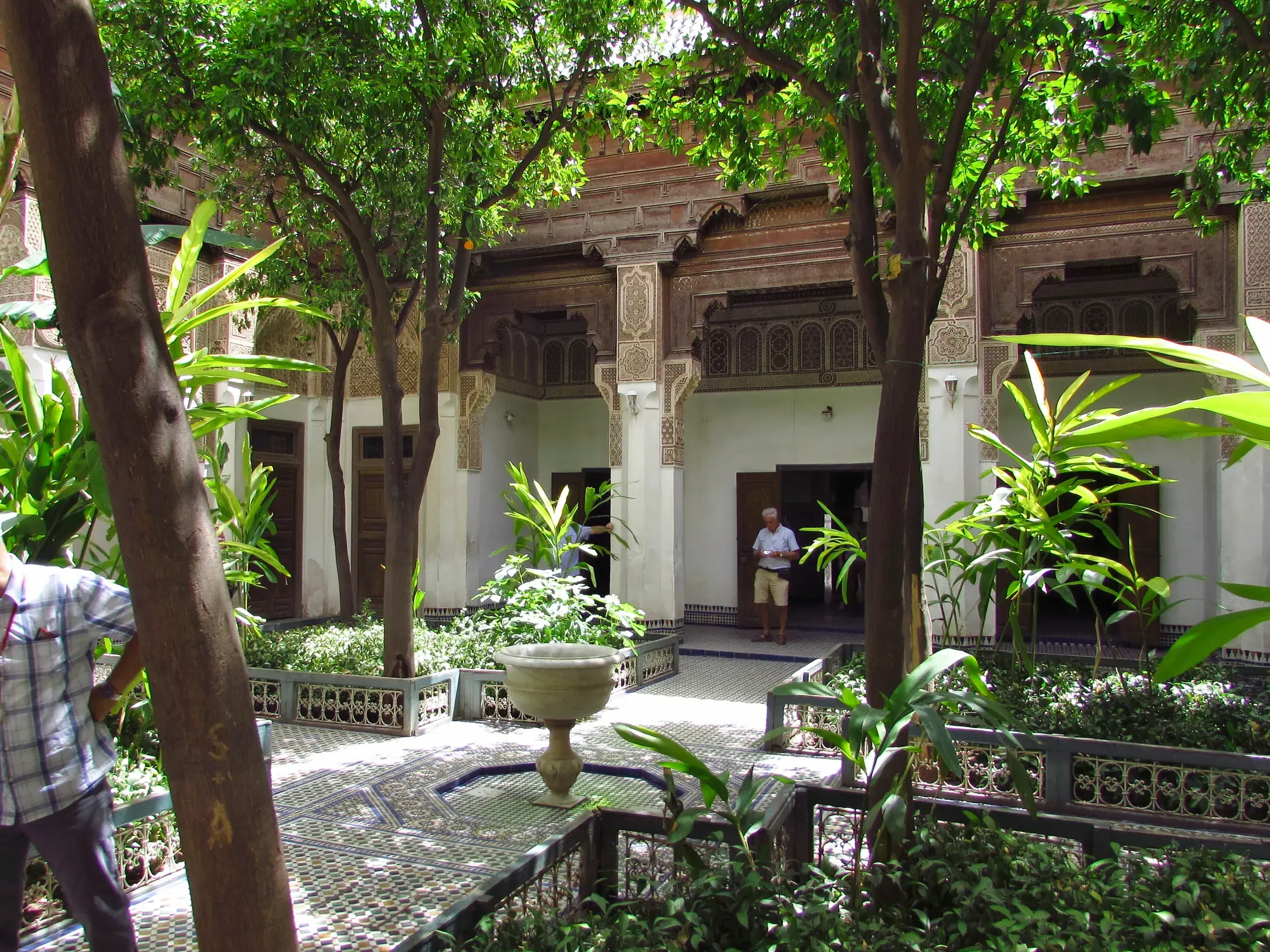 Ein Reiseleiter in einem grünen, sonnigen Innenhof des Bahia-Palasts in Marrakesch mit Mosaiken und Holzschnitzereien.