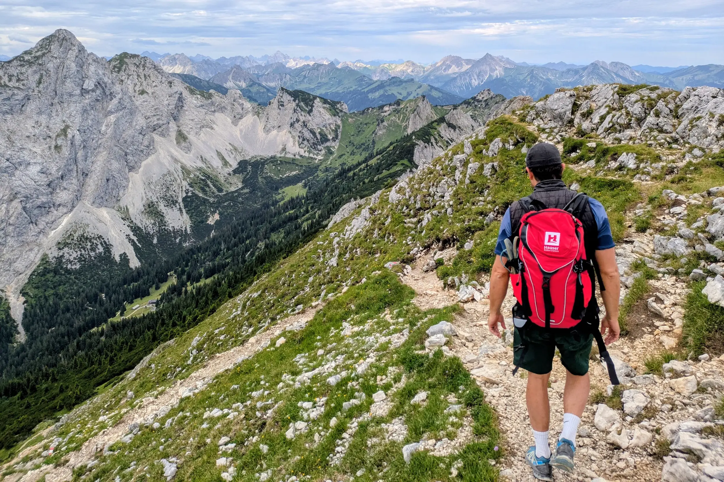 Ein Wanderer mit rotem Rucksack auf einem Grat in den Ammergauer Alpen mit Panoramablick auf die Gipfel des Ostallgäus.