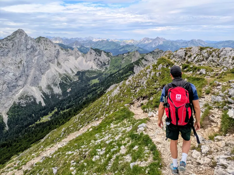 Ein Wanderer mit rotem Rucksack auf einem Grat in den Ammergauer Alpen mit Panoramablick auf die Gipfel des Ostallgäus.