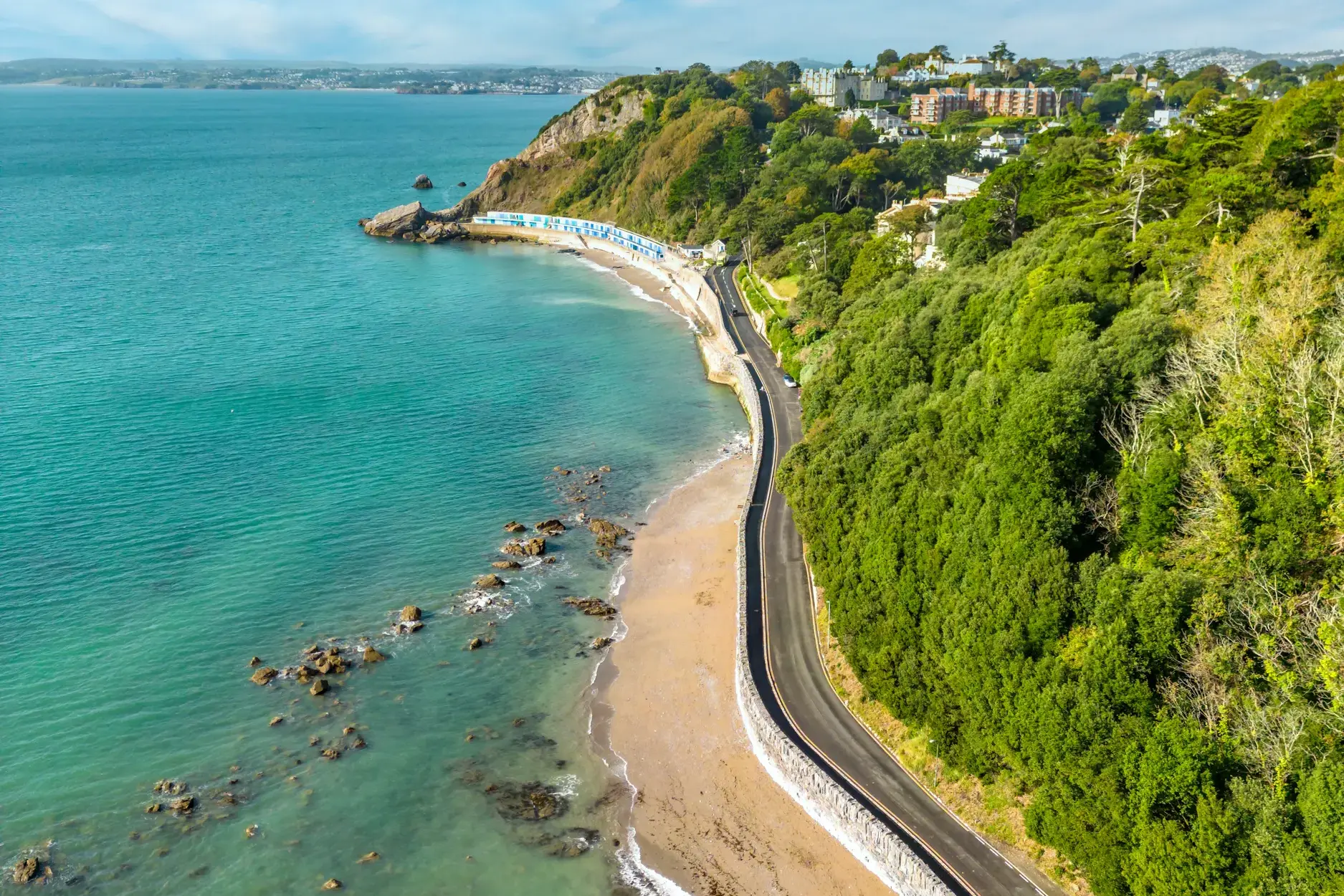 Luftaufnahme der Englischen Riviera bei Paignton: eine Küstenstraße führt entlang des türkisen Meeres und bewaldeter Hänge.