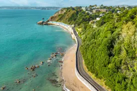 Luftaufnahme der Englischen Riviera bei Paignton: eine Küstenstraße führt entlang des türkisen Meeres und bewaldeter Hänge.