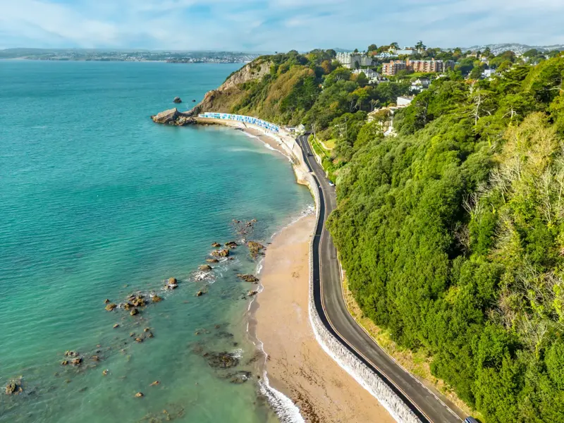 Luftaufnahme der Englischen Riviera bei Paignton: eine Küstenstraße führt entlang des türkisen Meeres und bewaldeter Hänge.