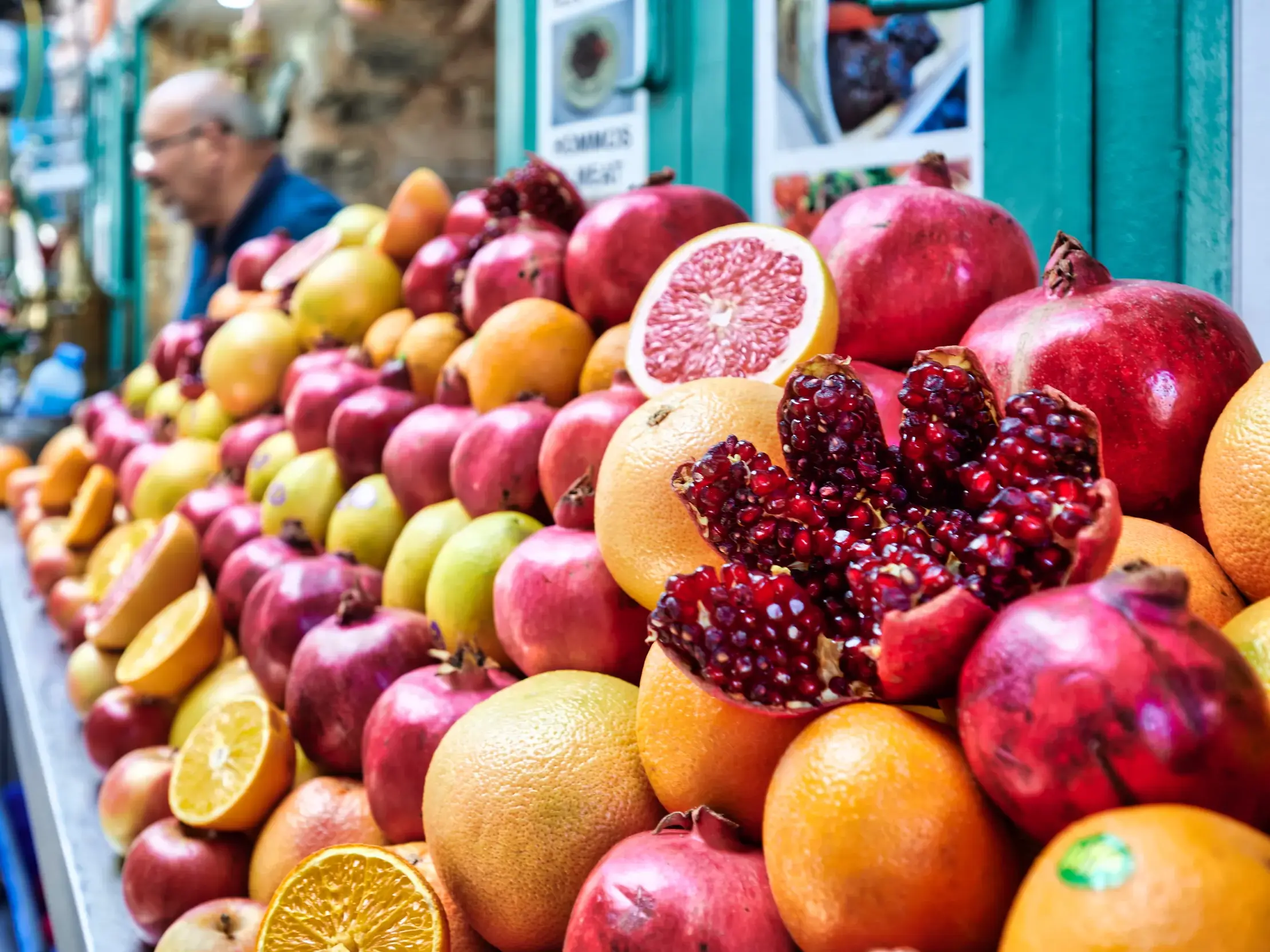 Bunter Marktstand mit aufgetürmten Granatäpfeln und Orangen auf einem Basar in Jerusalem, ein Verkäufer im Hintergrund.